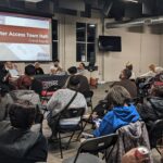 A town hall meeting with a "Voter Access Town Hall: Grand Rapids" presentation on a screen and panelists seated at a table in front of an audience.